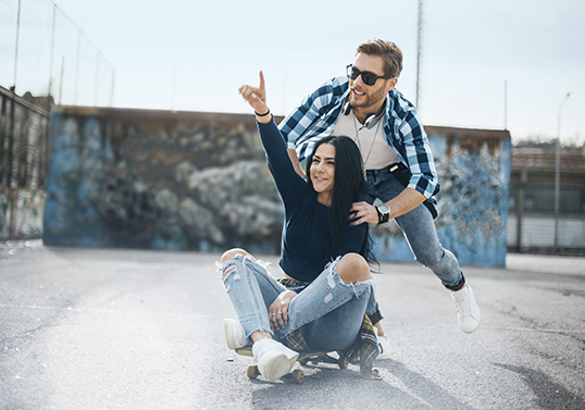 Ein junger Mann mit Sonnenbrille schiebt eine junge Frau, die auf einem Skateboard sitzt. Sie streckt fröhlich den Arm in die Luft und zeigt etwas mit ihrem Zeigefinger.